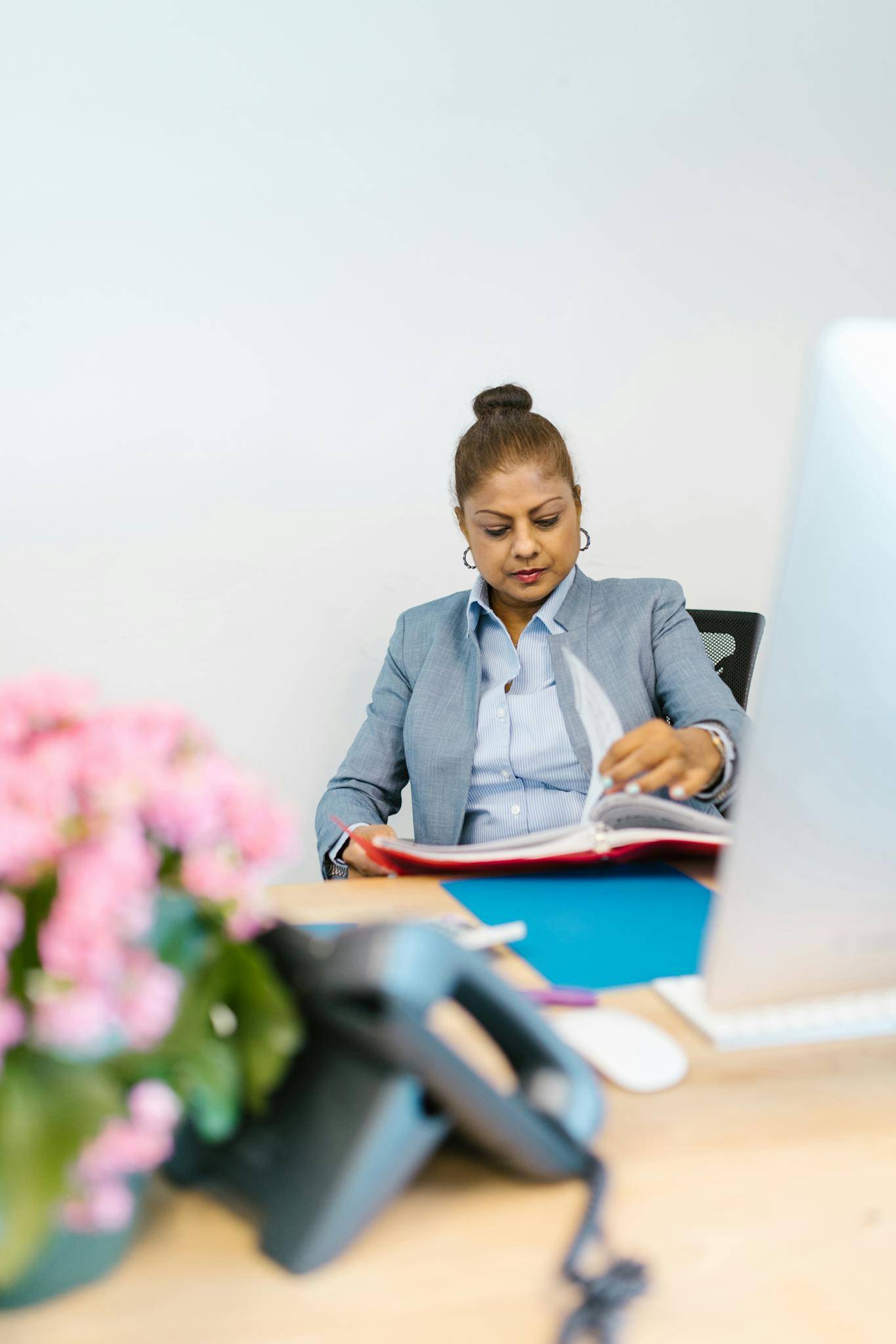Confident businesswoman in office reading documents at desk with flowers and computer.